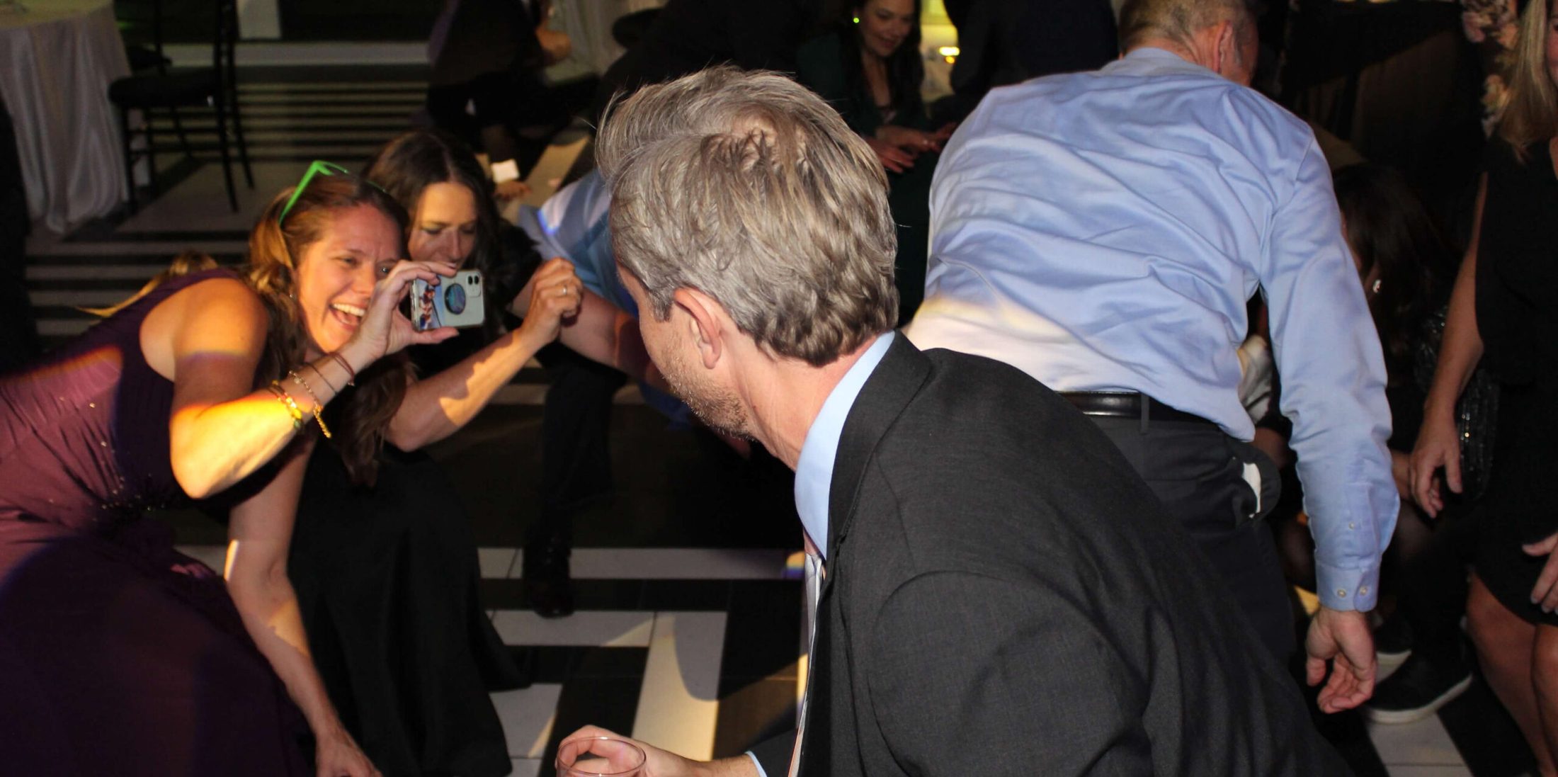 Two women bent down with a camera pointed at a man holding a drink, dancing on the dance floor with Paul Michaels Events DJ Company playing music