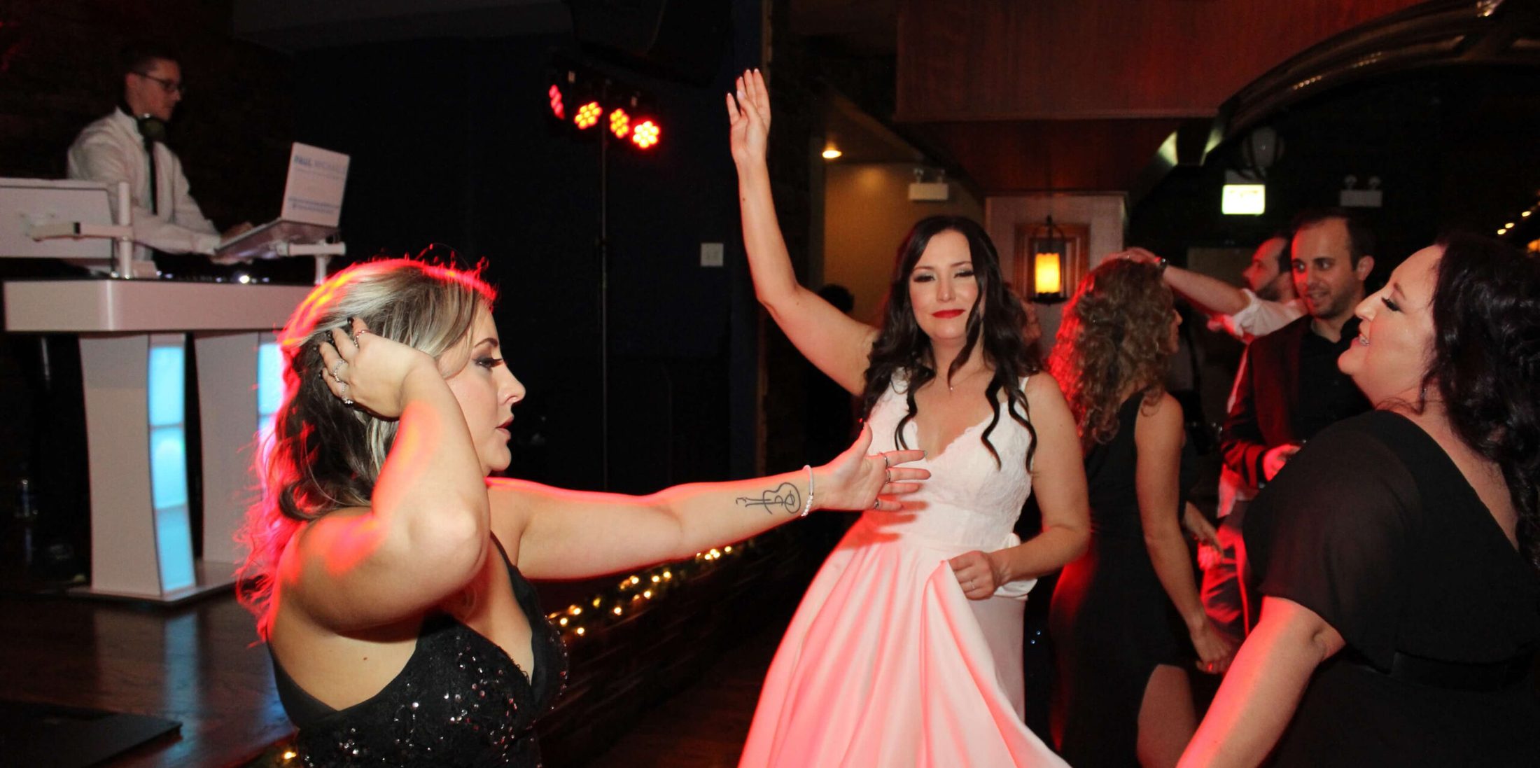 Photo of three women on the dance floor - one doing the sprinkler, one (the bride) with her hands in the air, and the third moving her hips. In the background, you can see DJ Tyler from Paul Michaels Events DJ Company playing the best music.