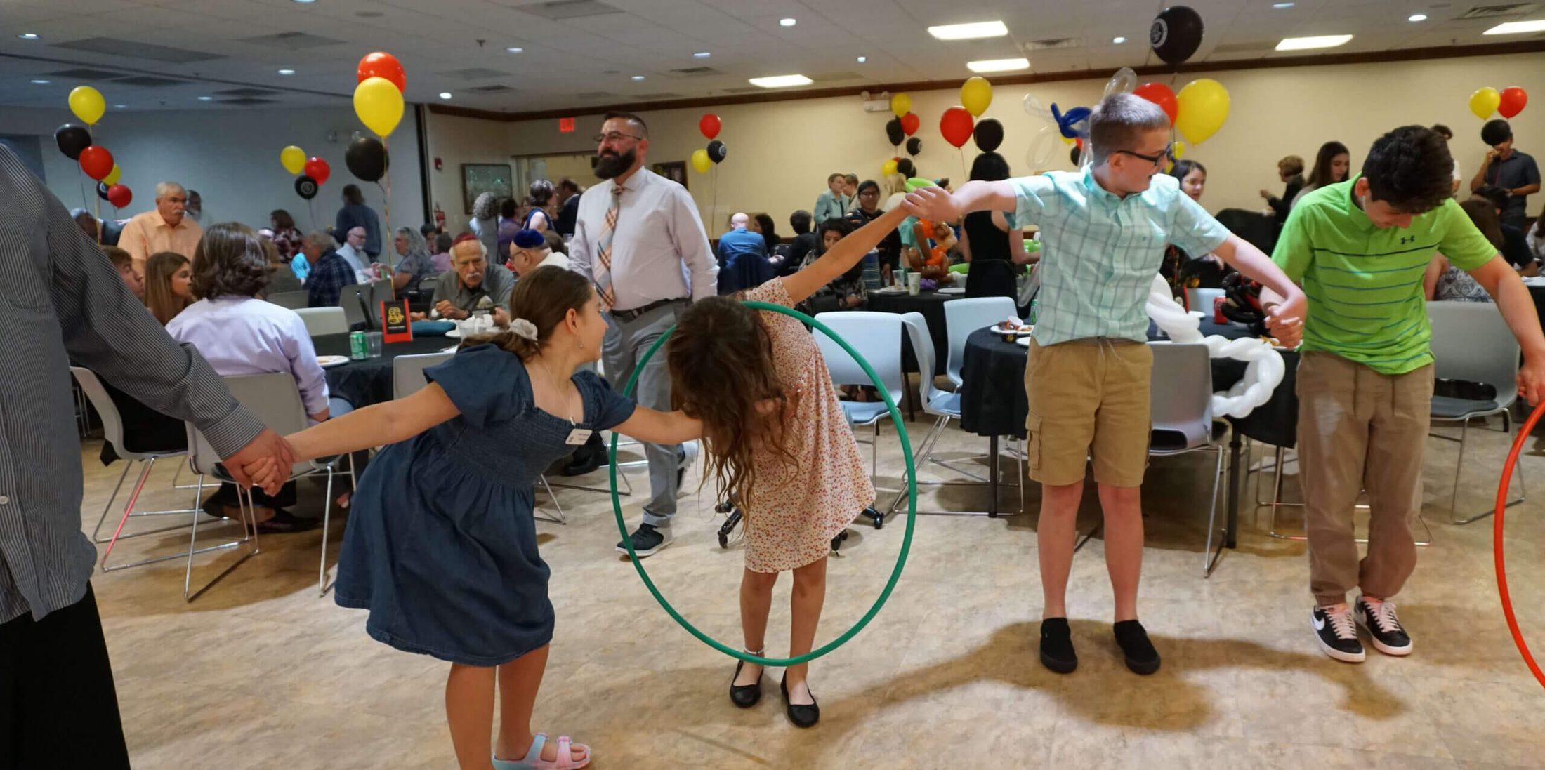 A group of kids holding hands in a chain link and passing a hula hoop to each other using their bodies, with entertainment being provided by Paul Michaels Events, an expert in B'nai Mitzvah entertainment services